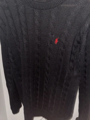 Svart kabelstickad tröja Polo Ralph Lauren - Kabelstickad svart tröja från Polo Ralph Lauren med rund halsringning och röd broderad logga på bröstet. Klassisk och tidlös design i mjukt material, perfekt för lager-på-lager och snygg till jeans.