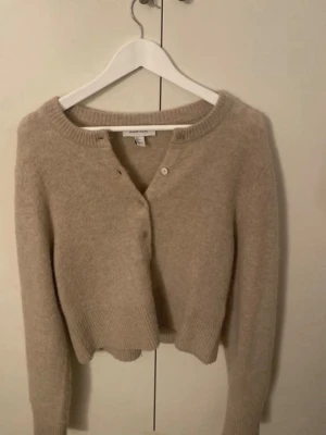 Beige kofta från other stories  - Mysig beige kofta från other stories  med rund halsringning och långa ärmar. Koftan har en croppad passform och stängs med knappar framtill. Tillverkad i ett mjukt material som känns skönt mot huden.