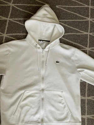 Vit zip hoodie från Lacoste - Snygg vit hoodie från Lacoste med dragkedja framtill och klassisk krokodil-logga på bröstet. Hoodien har justerbar huva med snören och två stora fickor framtill. Tillverkad i mjukt bomullsmaterial som är skönt att bära.