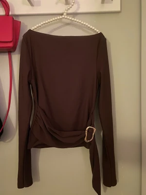 Brun omlottopp med spänne - Snygg brun omlottopp med lång ärm och dekorativt bälte i midjan med guldfärgat spänne. Toppen har en figurnära passform och båtringad hals. Perfekt att styla med jeans eller kjol för en trendig look. Använd 1 gång! 