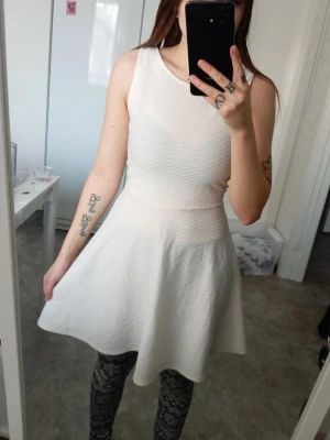 Vit ärmlös kortklänning med öppen rygg - Säljer en vit ärmlös kortklänning med strukturmönster och v-formad öppen rygg. Klänningen har en figurnära passning upptill och en utsvängd kjol. Perfekt för dig som gillar stilrena och enkla plagg.