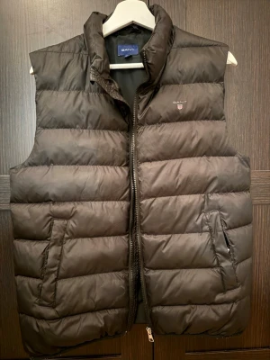 Svart dunväst från GANT - Snygg svart dunväst från GANT med hög krage och dragkedja framtill. Västen har två snedställda fickor med dragkedja och diskret GANT-logga på bröstet. Klassisk quiltad design och fyllning som håller värmen. Perfekt lager på lager-plagg för kyliga dagar.