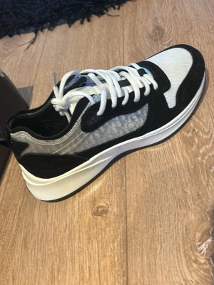 Dior b25 - Snygga sneakers från Dior i svart och vitt med grå detaljer. Ovandelen är i mesh och mocka, med vita skosnören och en chunky vit sula. Sidan har diskret Dior-mönster och sulan är märkt med Dior-loggan. Perfekta för dig som gillar exklusiv streetwear.