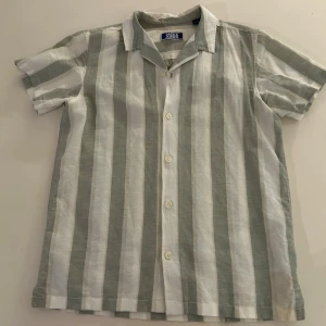 Randig skjorta från Jack & Jones - Snygg skjorta från Jack & Jones med breda vertikala ränder i vitt och ljusgrått. Skjortan har klassisk krage, knäppning framtill och korta ärmar. Perfekt för en avslappnad och trendig stil.