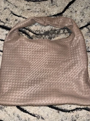 Beige flätad handväska - Snygg beige handväska från Gina Tricot med flätad design och mjukt konstläder. Väskan har ett rymligt huvudfack och magnetknäppning. Perfekt för dig som gillar trendiga accessoarer med stilren look.