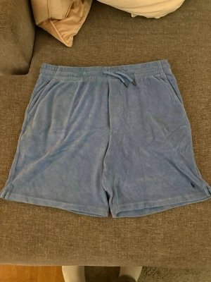 Blå frottéshorts från Polo Ralph Lauren - Mysiga blå shorts från Polo Ralph Lauren i mjuk frotté. De har snörning i midjan, sidofickor och en liten broderad logga nertill. Perfekta för chill dagar eller sommarhäng.
