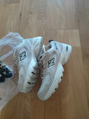 Vita New Balance 530 sneakers - Stilrena New Balance 530 sneakers i vitt med svarta och grå detaljer. Skorna har en chunky sula, meshpaneler för extra ventilation och klassisk N-logga på sidan. Perfekta för dig som gillar retrovibbar och sportig look.