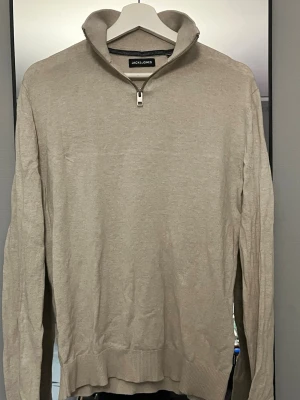 Beige halvzip tröja från Jack & Jones - Snygg beige tröja från Jack & Jones med halvzip och hög krage. Tillverkad i mjukt material med ribbade muddar vid ärmslut och nederkant. Perfekt för en avslappnad men stilren look.