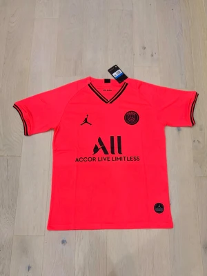 PSG x Jordan neonröd matchtröja M - Säljer en neonröd PSG x Jordan matchtröja i storlek M. Tröjan har svarta detaljer, PSG-logga och Jordan-logga på bröstet samt svart text 'Accor Live Limitless' framtill. Tillverkad i lätt och ventilerande material, perfekt för fotboll eller träning.