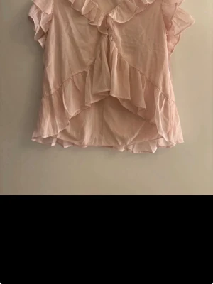 Ljusrosa volangblus från Nelly oanvänd  - Supersöt ljusrosa blus från Nelly med volangdetaljer och v-ringning. Blusen har korta ärmar och ett flowy, luftigt tyg som ger en romantisk vibe. Perfekt att styla med jeans eller kjol för en trendig look. Oanvänd 