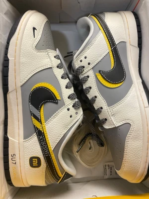 Nike x Xiaomi grå och vita sneakers - Snygga sneakers från Nike i samarbete med Xiaomi. Skorna har en grå och vit ovandel i läder med svarta och gula detaljer, inklusive en dubbellagd Swoosh. Svart och vitmönstrade skosnören och svart yttersula. Perfekta för dig som gillar unika collabs och streetwear.