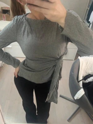 topp - Snygg grå långärmad topp med diskret rutigt mönster och knyt i midjan. Toppen har rund halsringning och är figurnära i modellen. Perfekt att styla med både jeans och kjol.