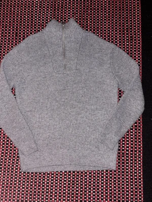 Grå stickad tröja med halv dragkedja - Snygg grå stickad tröja med ribbad struktur och halv dragkedja framtill. Tröjan har hög krage och långa ärmar, perfekt för kyliga dagar. Enkel och stilren design som passar till det mesta.