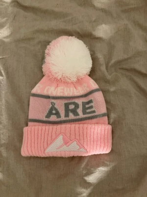 Åre mössa - Säljer en söt rosa mössa från Cheapo med stor fluffig tofs i vitt och grått. Mössan har texten 'ÅRE' framtill och ett broderat bergmotiv på den ribbstickade uppvikta kanten. Perfekt för kalla dagar och snygg till vinterlooken.