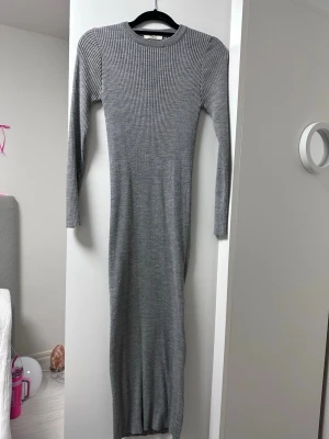 Grå ribbad långklänning - Snygg grå långklänning i ribbad bomullsmix med lång ärm och rund hals. Klänningen har en kroppsnära passform och är perfekt för dig som gillar en enkel men stilren look.