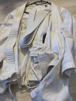 Vit karate-gi med bälte  - Säljer en klassisk vit karate-gi från Budo-Nord med tillhörande vitt bälte. Dräkten består av jacka och byxor med dragsko i midjan. Tillverkad i slitstarkt bomullstyg och har förstärkta sömmar för extra hållbarhet. Perfekt för träning eller tävling.