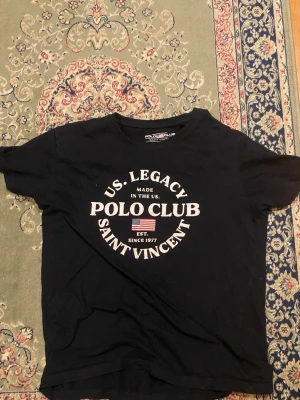Svart Polo Club t-shirt med tryck - Cool svart t-shirt från Polo Club med stort vitt tryck på bröstet och amerikansk flagga. Klassisk rund hals och korta ärmar, perfekt för en avslappnad look. Materialet är mjukt och troligtvis bomull. Passar dig som gillar streetstyle och tydliga loggor.