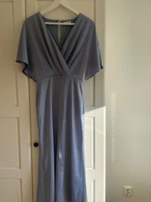 Ljusblå långklänning - Elegant ljusblå långklänning från NA-KD med omlott framtill och v-ringad hals. Klänningen har vida, korta ärmar och ett mjukt, flytande fall. Perfekt för sommarens alla tillfällen. Passar 34/36