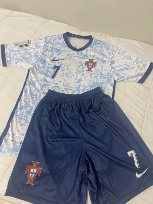 Portugal fotbollskit Nike blå/vit - Snyggt Portugal fotbollskit från Nike med matchtröja i vitt och ljusblått mönster samt blå shorts. Båda delarna har nummer 7 och Portugals landslagsmärke. Tröjan har rund hals och korta ärmar, shortsen har resår i midjan. Perfekt för fotbollsträning eller match. Jätte fin ronaldo set, knappt andvänd tror de är storlek s/xs. Köp bundell så får du rabbat! 
