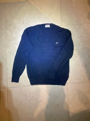 Mörkblå stickad tröja från Gant - Klassisk mörkblå stickad tröja från Gant med V-ringning och diskret broderad logga på bröstet. Tröjan har långa ärmar och ribbade muddar vid ärmslut och nederkant. Perfekt för lager på lager och enkel att matcha.