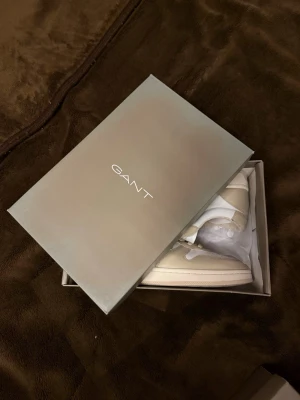 Beige och vita sneakers från Gant - Snygga sneakers från Gant i beige och vitt med broderad logga på sidan. Skorna har en klassisk låg siluett, vita snören och detaljer i mocka och läder. Perfekta för dig som vill ha en clean och stilren look. Helt ny