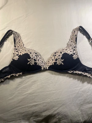 Mörkblå spets-bh med beige detaljer - Säljer snygg marinblå bh med vadderade kupor och dekorativa beige spetsdetaljer längs kanterna och axelbanden. Bh:n har justerbara axelband och hakknäppning bak. Säljer då den är för liten🫶🫶