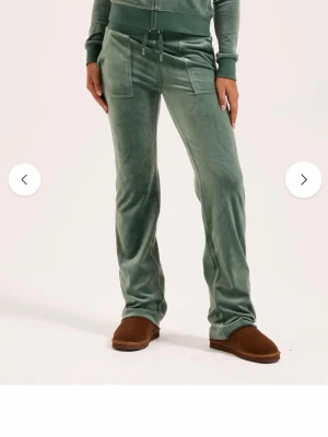 Gröna velourbyxor från Juicy Couture - Mjukisbyxor i grön velour från Juicy Couture med raka ben, bred mudd i midjan och snörning. Byxorna har stora fickor fram och bak med lock och silverfärgade knappar. Använda få tal gånger och i perfekt skick