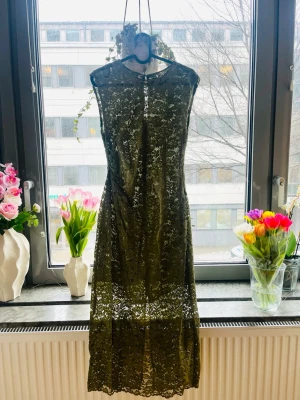 Olivgrön spetsklänning med blommönster - Säljer en olivgrön långklänning i spets med blommigt mönster. Klänningen är ärmlös och har en elegant, rak passform. Perfekt för dig som vill ha en unik och stilren look. Materialet är spets och klänningen har en transparent känsla.