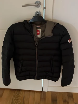 Svart pufferjacka från Colmar - Snygg svart pufferjacka från Colmar med huva och dragkedja framtill. Jackan har två sidofickor och Colmar-logga på ärmen. Perfekt för kalla vinterdagar och har en klassisk, puffig design. Materialet är quiltat och ger en sportig look.