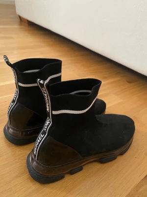 Svarta sock-sneakers från Michael Kors - Svarta sock-sneakers från Michael Kors med vit rand runt ankeln och logotyp på hälen. Ovandelen är i stretchigt tyg och sulan är chunky och svart med bruna detaljer. Snygg och modern design som sticker ut.