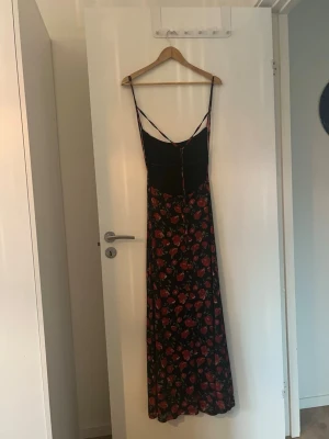 Svart långklänning med röda rosor - Långklänning med smala axelband och v-ringning. Klänningen är svart med ett blommigt mönster av röda rosor och gröna blad. Figurnära passning upptill och faller rakt ned. Perfekt för sommarens alla tillfällen.