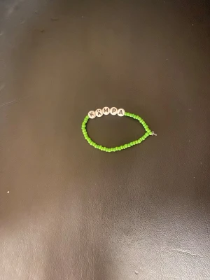 Grönt armband med bokstavspärlor - Armband med små gröna pärlor och vita runda bokstavspärlor som bildar ordet 'KÄMPA'. Trädd på elastisk tråd och har en enkel, ungdomlig design. Perfekt för dig som vill ha ett personligt smycke med peppande budskap.