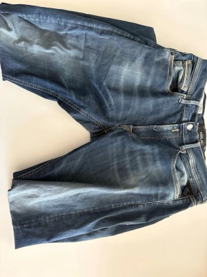 Blå Replay jeans med slitningar - Säljer ett par blå jeans från Replay med snygga slitningar och tvättade detaljer. Klassisk femficksmodell med raka ben och läderpatch bak i midjan. Jeansen har kontrastsömmar och är tillverkade i denim med en cool vintagekänsla. Har ett litet hål som är enkelfixat 