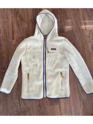 Patagonia fleece - Mysig vit fleecejacka från Patagonia med huva och hel dragkedja. Jackan har blå detaljer längs dragkedjan och fickor med snygga läderdragare. Tillverkad i fluffigt teddy-material som håller dig varm och bekväm. Perfekt för kyliga dagar och enkel att matcha med olika outfits.