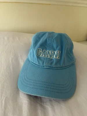 Blå keps från Ganni - Snygg ljusblå keps från Ganni med broderad vit logga framtill. Klassisk böjd skärm och justerbar rem baktill för perfekt passform. Tillverkad i bomullstyg som känns både bekvämt och stilrent. Perfekt accessoar för en avslappnad look.