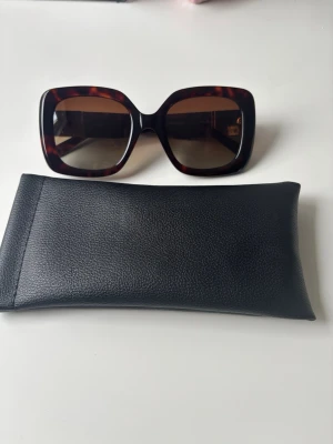 Bruna fyrkantiga solglasögon - Snygga solglasögon med breda, fyrkantiga bågar i brunsköldpaddsmönster. Glasen är mörka och bågarna har gulddetaljer vid gångjärnen. Märker är Molarache. Kommer med ett svart fodral i syntetmaterial. Perfekta för att lyfta din stil i solen. Helt oanvändna.