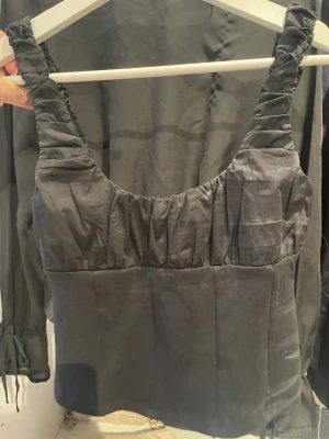 Svart linnetopp med rynkade axelband - Trendig svart linnetopp med breda, rynkade axelband och lätt glansigt tyg. Toppen har en rak passform och är perfekt för att styla med jeans eller kjol. Enkel men ändå snygg design som ger en cool vibe.