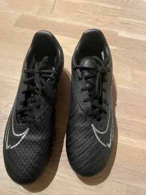 Svarta Nike fotbollsskor med snörning - Säljer ett par svarta fotbollsskor från Nike med klassisk snörning och vita Nike-loggor på ovansidan. Skorna har en låg profil och är tillverkade i syntetmaterial med texturerad yta för bättre bollkontroll. Perfekta för dig som vill ha stilrena och funktionella skor på planen.