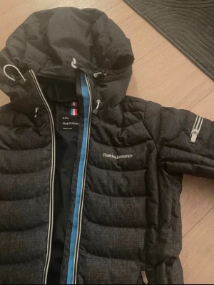 Mörkgrå jacka Peak Performance - Snygg Mörkgrå höst jacka från Peak Performance med huva och dragkedja framtill. Jackan har quiltad design, logga på bröstet och praktisk ficka med dragkedja på ärmen. Nyskick! 