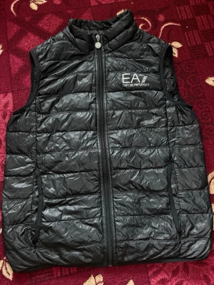 EA7 Emporio Armani vest - Säljer en stilren och exklusiv svart Emporio Armani väst i mycket bra skick. Västen är sparsamt använd och har inga skador, fläckar eller slitningar. Den har en modern och tidlös design som passar perfekt både till vardags och mer uppklätt. Lätt och bekväm att bära, perfekt över hoodie, tröja eller långärmad.