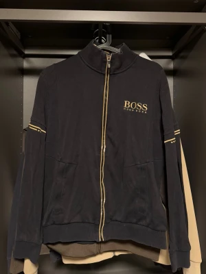 Svart zip hoodie från BOSS - Snygg svart hoodie från BOSS med guldfärgade detaljer och dragkedja framtill. Stort BOSS-tryck på bröstet och ribbade partier på ärmarna. Två fickor fram och sportig look. Perfekt för dig som gillar stilrena och exklusiva plagg.