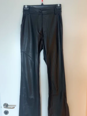 Svarta skinnbyxor med raka ben - Snygga svarta skinnbyxor med hög midja och raka ben. Byxorna har bälteshällor, knapp och dragkedja framtill. Från madlady