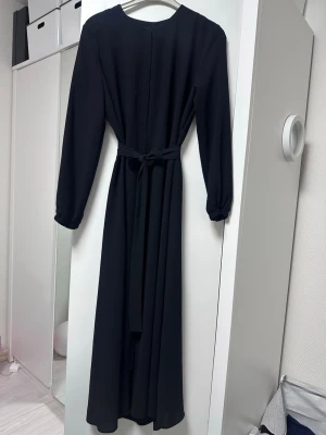 Svart långklänning med bälte  - Stilren svart långklänning med långa ärmar och ett matchande bälte i midjan. Klänningen har rund halsringning och ett enkelt, elegant fall. Perfekt för dig som gillar minimalistisk och klassisk stil.