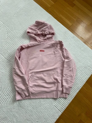 Ljusrosa hoodie från Levi's - Säljer en ljusrosa hoodie från Levi's med klassisk röd logga på bröstet. Hoodien har en stor känguruficka framtill, ribbade muddar och en skön huva. 