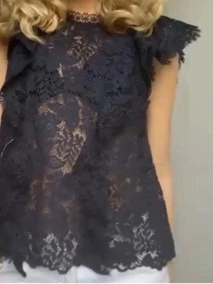 Svart spetsblus med volangdetaljer - Snygg svart blus i spets med blommigt mönster och volangdetaljer vid axlarna. Ärmlös modell med rund halsringning och transparent material som ger en trendig look. Perfekt att styla med jeans eller kjol för en cool outfit.