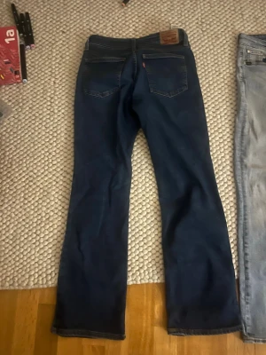 Mörkblå bootcut jeans från Levi's - Säljer ett par mörkblå Levi's jeans med klassisk bootcut-modell. Jeansen har fem fickor, läderpatch bak i midjan och subtila slitningar framtill för en snygg vintage-look. Tillverkade i slitstark denim med en tidlös passform.