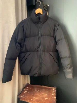 Svart pufferjacka/dunjacka med hög krage - Snygg och stilren mattsvart (ser svartare ut i verkligheten) pufferjacka med hög krage och dragkedja framtill. Jackan har en boxig passform och breda kanaler som ger en riktigt puffig look. Perfekt för kalla dagar.