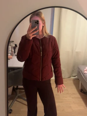 Vinröd pufferjacka med dragkedja - Snygg vinröd pufferjacka med quiltad design och dragkedja framtill. Jackan har en klassisk krage och långa ärmar, perfekt för våren. Passar dig som vill ha en stilren och bra jacka till våren. Storlek 40 skulle säga att de är en S. Aldrig använd.