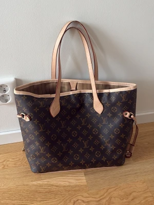  Monogram väska - Säljer en klassisk handväska i brunt monogramcanvas med beige läderdetaljer och långa handtag. Väskan har öppen topp, sidoremmar och randigt innerfoder. Perfekt för dig som gillar lyxig och tidlös design.