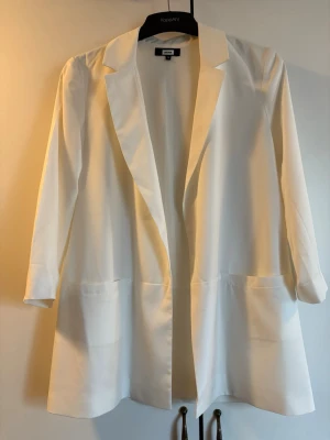 Vit kavaj från Bikbok - Stilren vit kavaj från Bikbok stl S med klassisk krage och långa ärmar. Modellen är rak och har två stora fickor framtill. Perfekt för en clean och modern look. Materialet känns mjukt och har en lätt struktur.